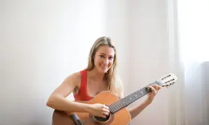 Lächelnde Frau mit blonden Haaren übt das Gitarre lernen für Erwachsene. Sie sitzt auf dem Boden und spielt auf einer Akustikgitarre.