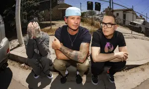 Die Mitglieder von blink-182 hocken auf dem Bürgersteig. Links Travis Barker, der sein Gesicht verdeckt. In der Mitte Mark Hoppus mit Cap und tätowierten Armen. Rechts Tom DeLonge mit Brille und Irokesenschnitt.
