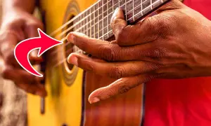 Hände spielen Fingerstyle Gitarre. Ein roter Pfeil zeigt auf die Finger, die die Saiten greifen.