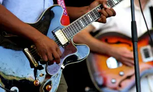 Hände spielen auf einer schwarzen E-Gitarre, während im Hintergrund eine weitere Gitarre zu sehen ist. Die Band probt für ihren nächsten Auftritt.