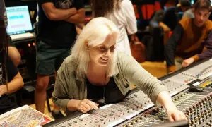 Sylvia Massy, lächelnd, bedient ein Mischpult in einem Tonstudio. Menschen im Hintergrund sind beschäftigt.