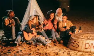 Freunde sitzen am Strand um ein Feuer, spielen Gitarre und singen Lagerfeuerlieder zum Mitsingen. Ein Zelt mit Lichterkette im Hintergrund.