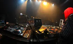 DJ-Pult mit Plattenspielern, Laptop und Mikrofon. Im Hintergrund eine Bühne mit Scheinwerfern und Publikum. Die DJ Namen sind hier nicht sichtbar.