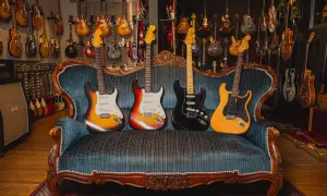 Vier E-Gitarren in verschiedenen gitarren formen und Farben ruhen auf einer antiken Couch, umgeben von einer Wand voller weiterer Gitarren.
