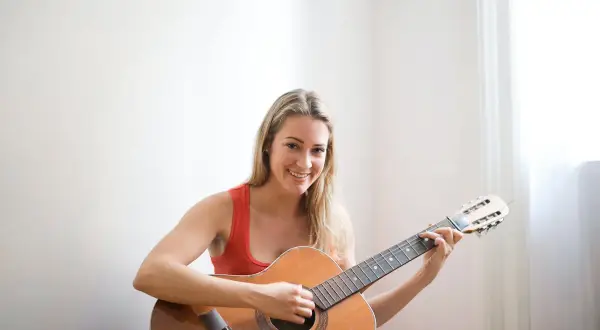 Lächelnde Frau mit blonden Haaren übt das Gitarre lernen für Erwachsene. Sie sitzt auf dem Boden und spielt auf einer Akustikgitarre.