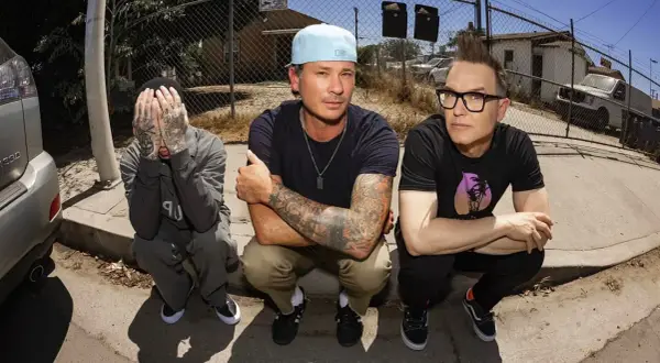 Die Mitglieder von blink-182 hocken auf dem Bürgersteig. Links Travis Barker, der sein Gesicht verdeckt. In der Mitte Mark Hoppus mit Cap und tätowierten Armen. Rechts Tom DeLonge mit Brille und Irokesenschnitt.