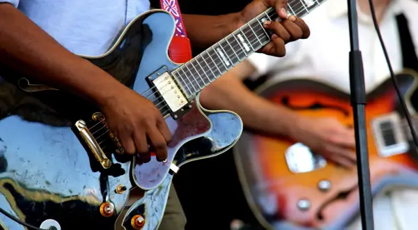 Hände spielen auf einer schwarzen E-Gitarre, während im Hintergrund eine weitere Gitarre zu sehen ist. Die Band probt für ihren nächsten Auftritt.