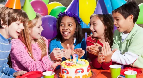 Kinder mit bunten Partyhüten singen Geburtstagslieder für ein Mädchen mit einem Kuchen.