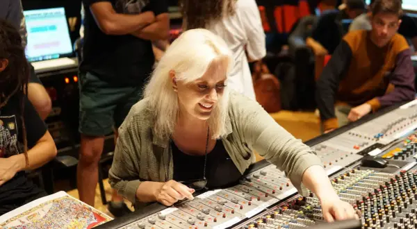 Sylvia Massy, lächelnd, bedient ein Mischpult in einem Tonstudio. Menschen im Hintergrund sind beschäftigt.