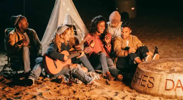 Freunde sitzen am Strand um ein Feuer, spielen Gitarre und singen Lagerfeuerlieder zum Mitsingen. Ein Zelt mit Lichterkette im Hintergrund.