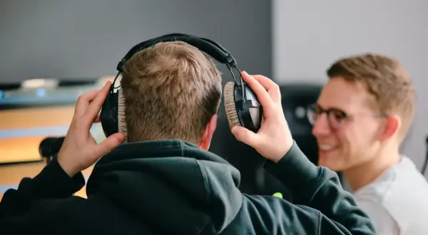 Zwei Männer bereiten sich darauf vor, einen Podcast aufzunehmen. Einer setzt sich Kopfhörer auf, während der andere lächelnd zusieht.