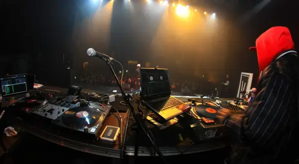 DJ-Pult mit Plattenspielern, Laptop und Mikrofon. Im Hintergrund eine Bühne mit Scheinwerfern und Publikum. Die DJ Namen sind hier nicht sichtbar.