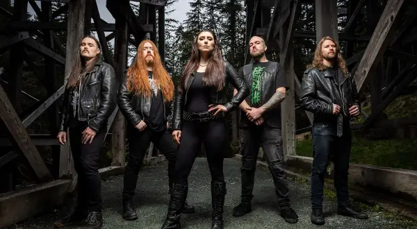 Fünf Musiker in Lederjacken, die an Metal Bands der 80er erinnern, posieren vor einer Holzkonstruktion im Wald.
