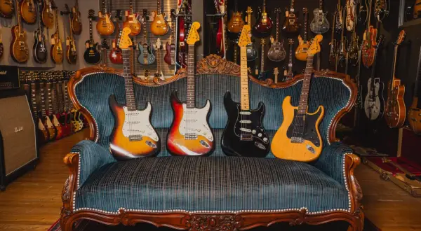 Vier E-Gitarren in verschiedenen gitarren formen und Farben ruhen auf einer antiken Couch, umgeben von einer Wand voller weiterer Gitarren.