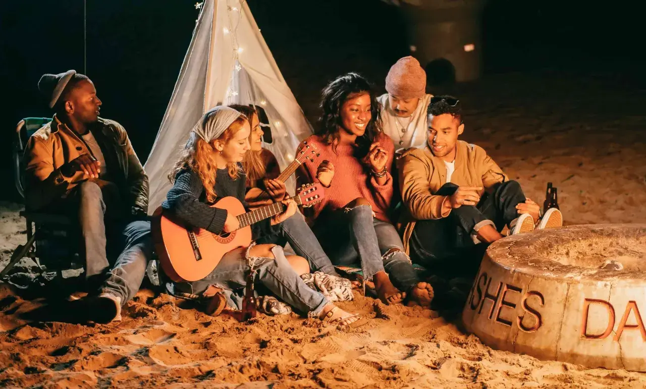 Freunde sitzen am Strand um ein Feuer, spielen Gitarre und singen Lagerfeuerlieder zum Mitsingen. Ein Zelt mit Lichterkette im Hintergrund.