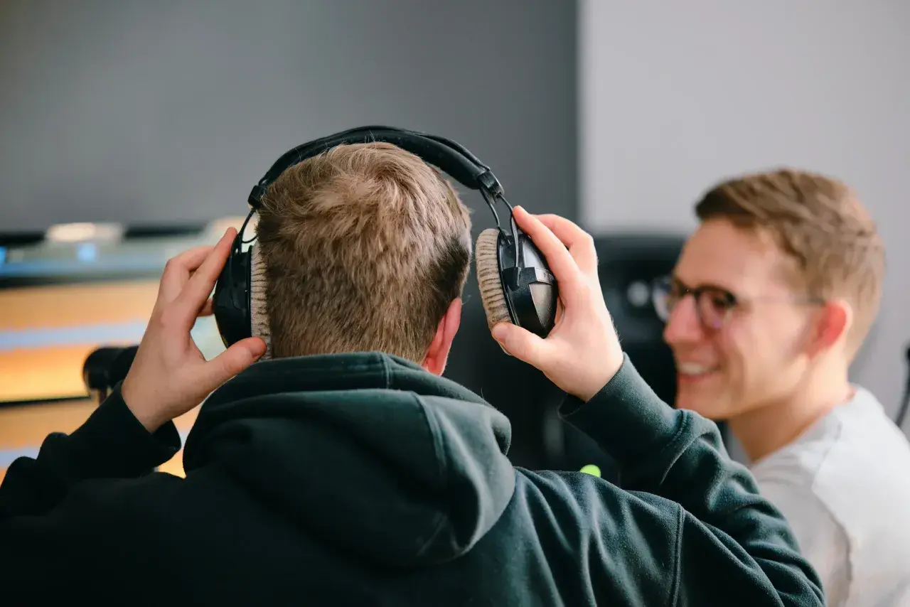 Zwei Männer bereiten sich darauf vor, einen Podcast aufzunehmen. Einer setzt sich Kopfhörer auf, während der andere lächelnd zusieht.
