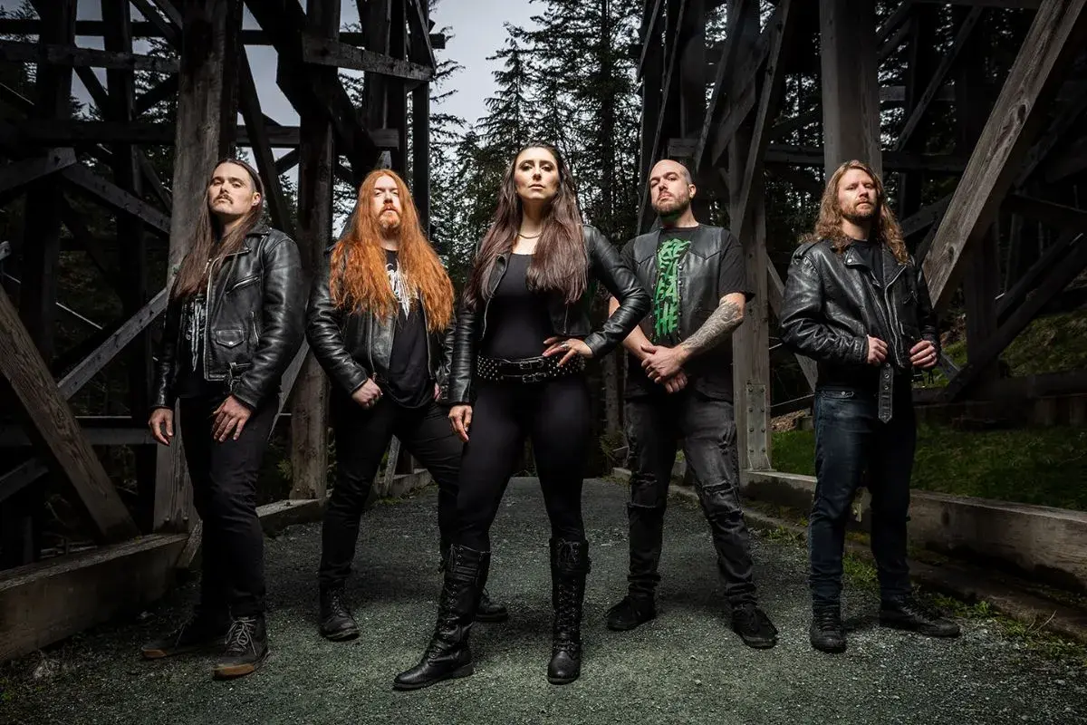 Fünf Musiker in Lederjacken, die an Metal Bands der 80er erinnern, posieren vor einer Holzkonstruktion im Wald.