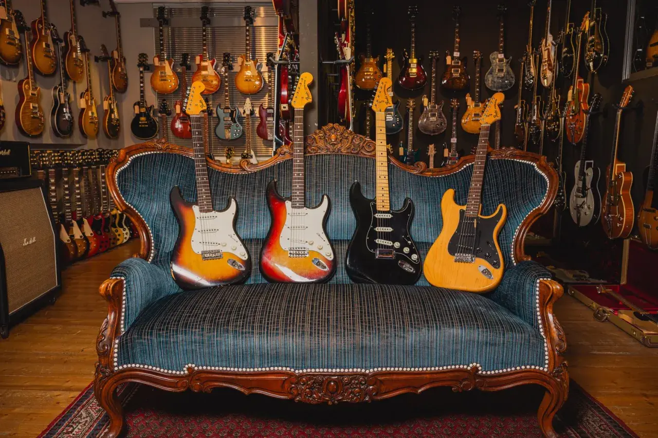 Vier E-Gitarren in verschiedenen gitarren formen und Farben ruhen auf einer antiken Couch, umgeben von einer Wand voller weiterer Gitarren.