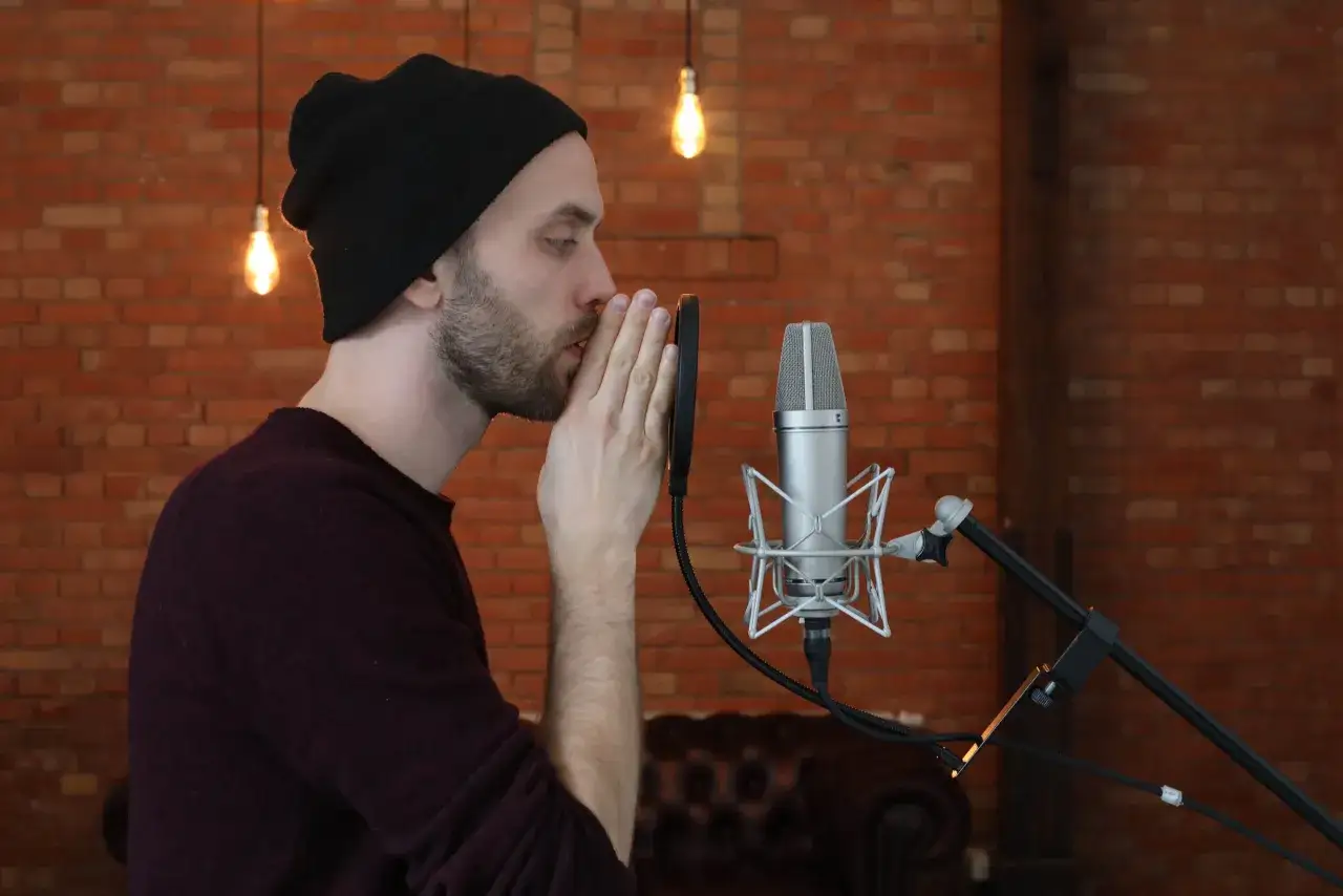 Mann mit schwarzer Mütze spricht ins Mikrofon, sucht nach rap ideen. Ziegelwand und Glühbirnen im Hintergrund.