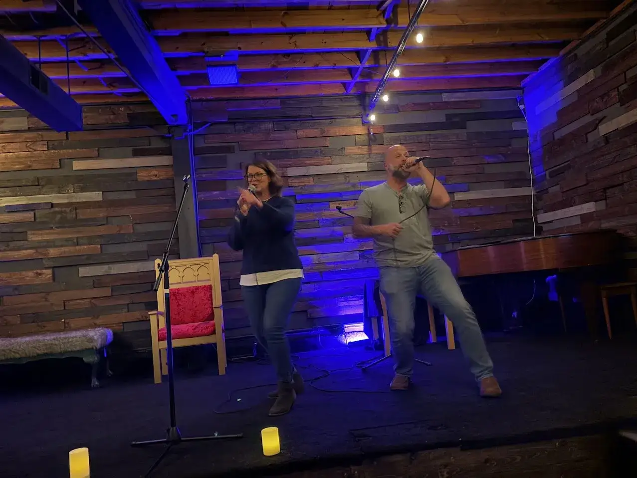 Ein Mann und eine Frau singen im Duett, vielleicht die top 100 karaoke songs. Sie stehen auf einer Bühne mit Holzwänden und blauer Beleuchtung.