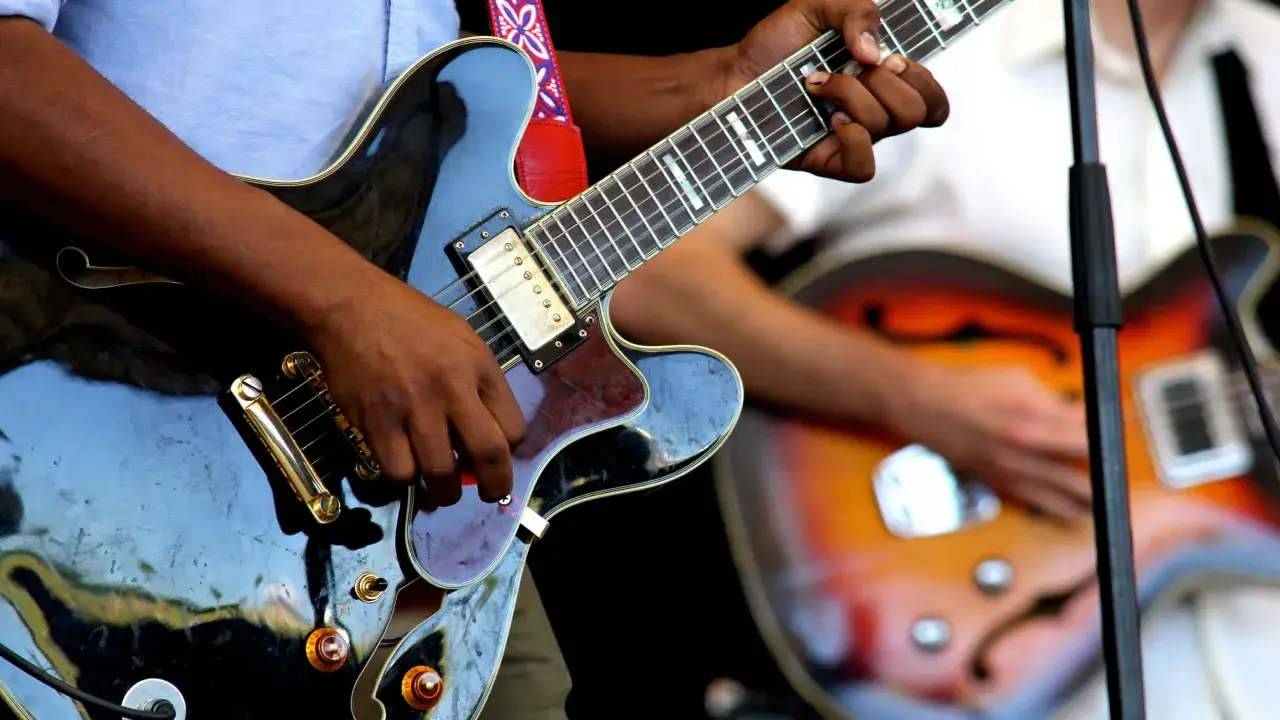 Hände spielen Gitarre, während im Hintergrund eine weitere Gitarre und ein Mikrofonständer zu sehen sind. Die Musik ist so gut, dass man fast die Band Namen erraten kann.