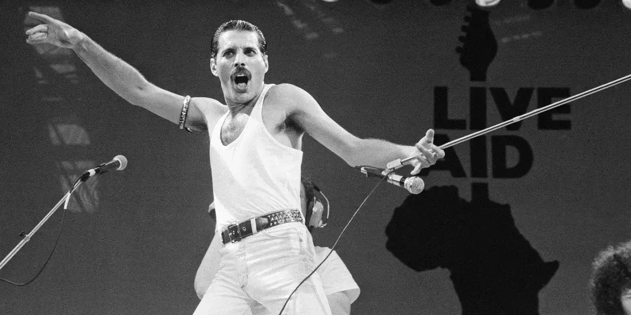 Freddie Mercury von der Gruppe Queen singt mit ausgestrecktem Arm und Mikrofon auf der Bühne.