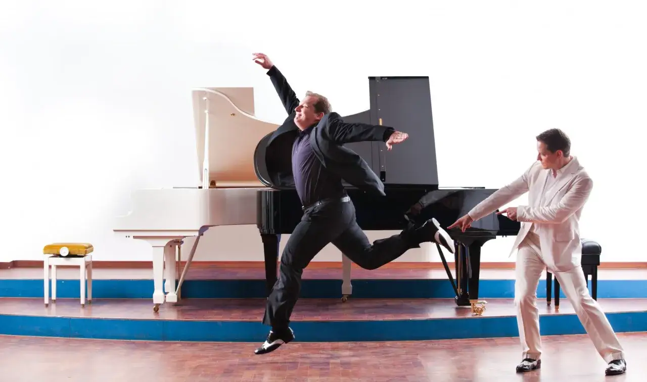 Ein Mann in Schwarz springt vor einem Flügel, während ein Mann in Weiß auf die Tasten zeigt. Vielleicht der beste Pianist der Welt in Aktion.