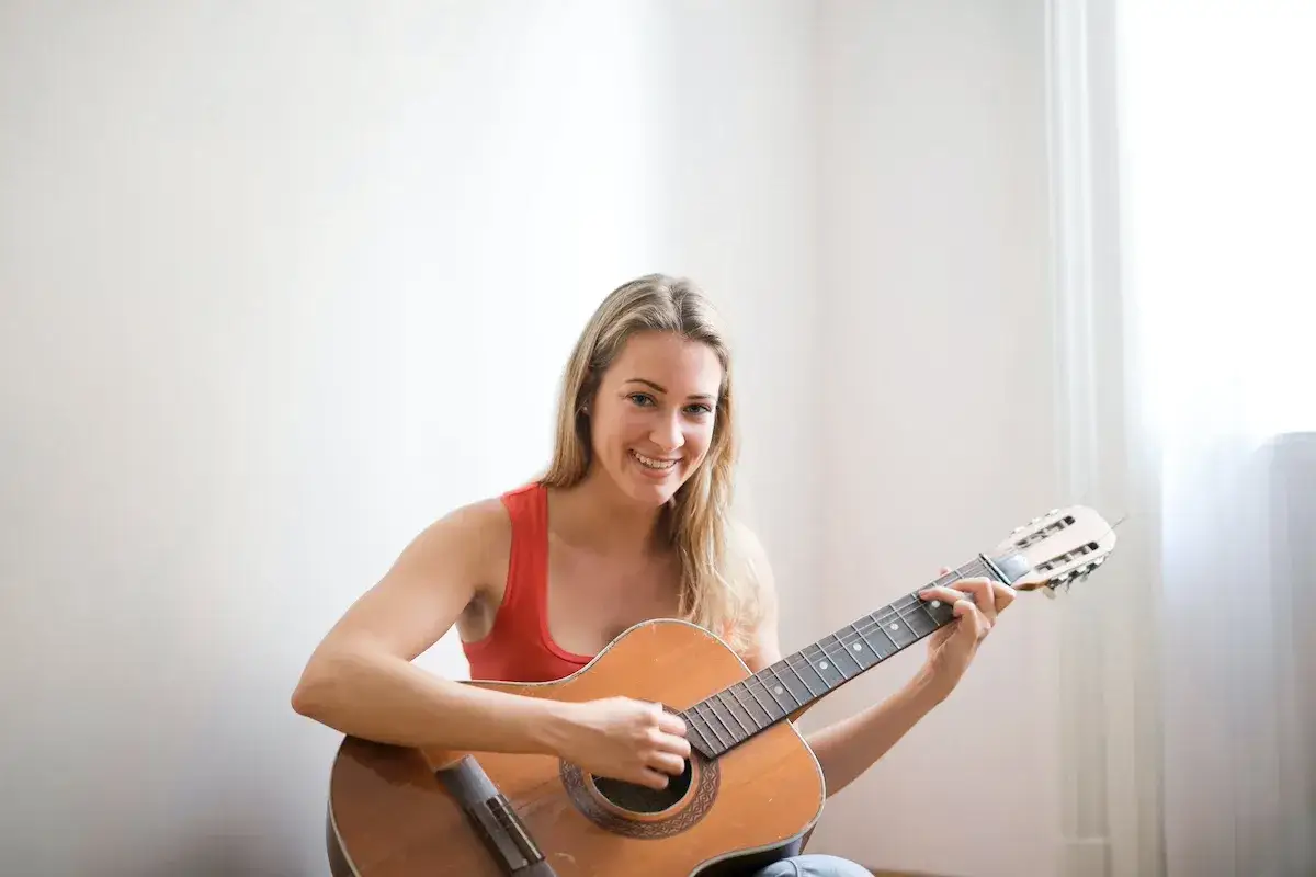 Lächelnde Frau mit blonden Haaren übt Gitarre spielen. Sie sitzt auf dem Boden und hält eine Akustikgitarre. Perfekt für alle, die Gitarre lernen erwachsene möchten.