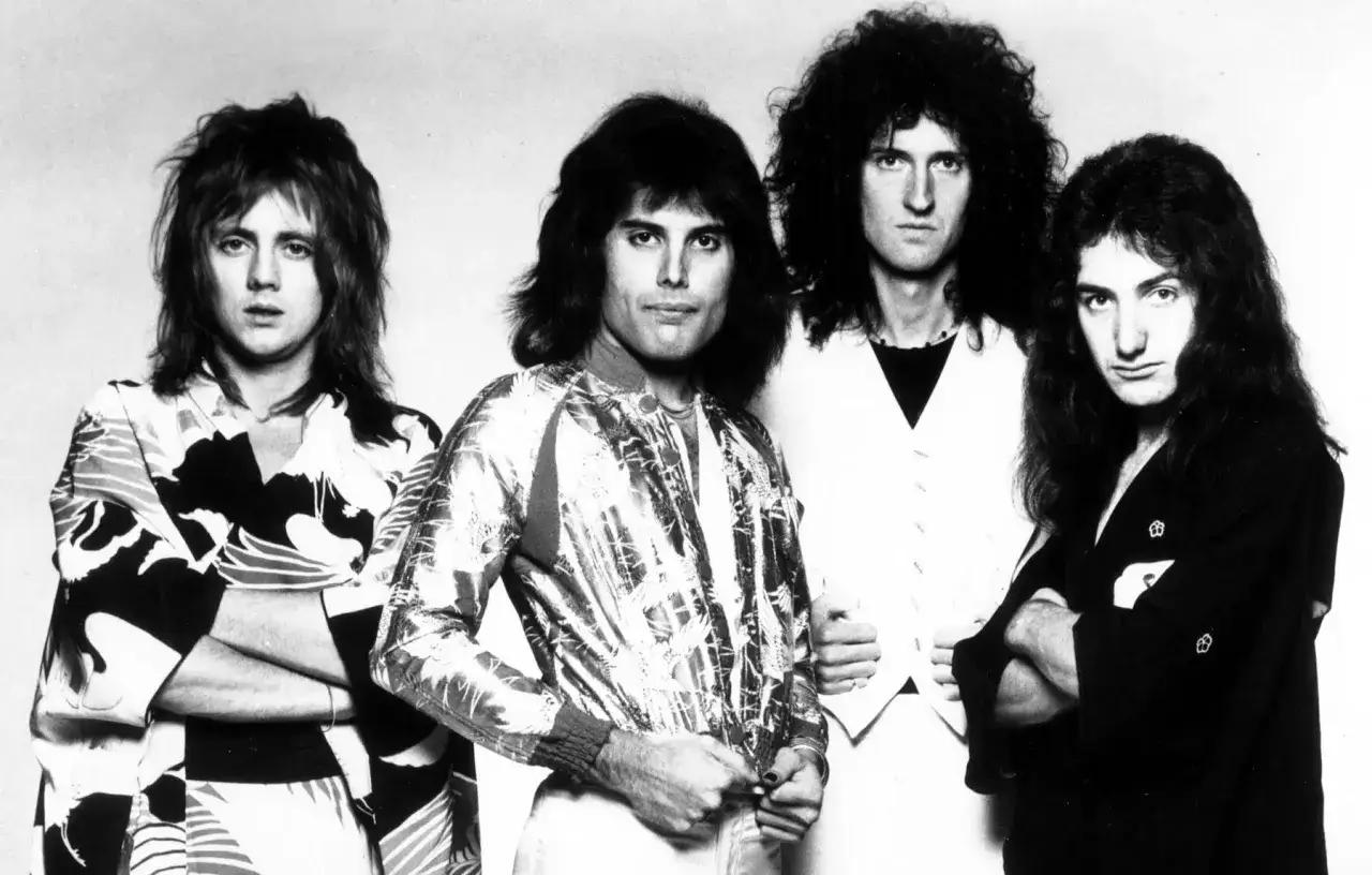 Die Mitglieder von Queen posieren für ein Schwarz-Weiß-Foto. Freddie Mercury trägt eine glänzende Jacke, Brian May eine Weste, Roger Taylor ein gemustertes Hemd und John Deacon einen dunklen Kimono.