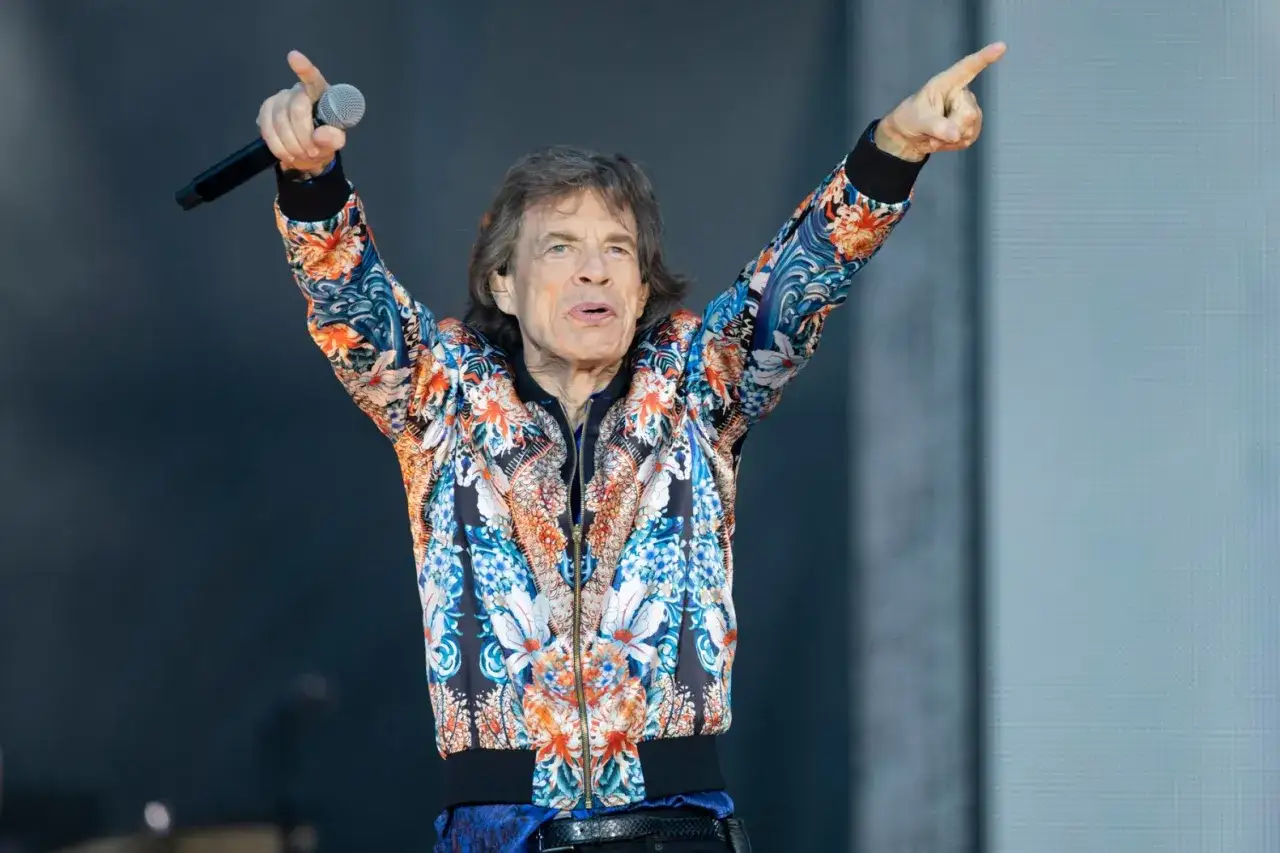 Mick Jagger, Frontmann der Mick Jagger Band, singt mit Mikrofon und erhobenen Armen in einer bunten Jacke.