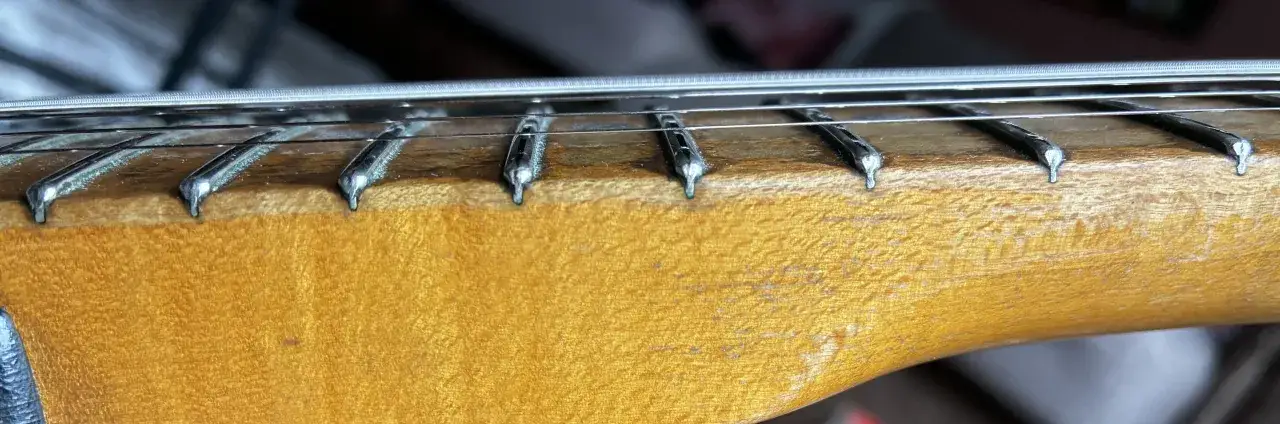 Nahaufnahme des Halses einer E-Gitarre mit Saiten und Bundstäbchen. Das Holz hat eine warme, honigfarbene Maserung.