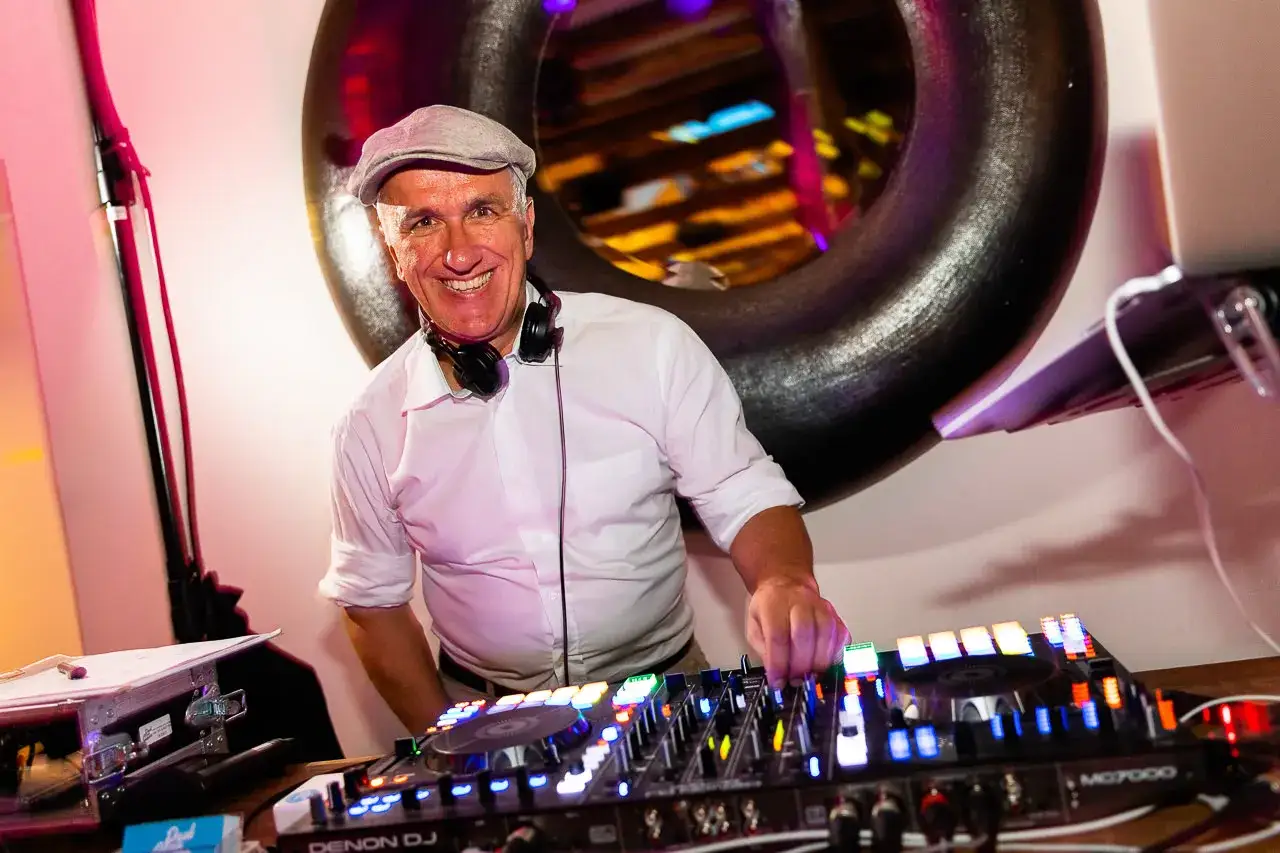 Ein DJ mit Kopfh&ouml;rern und Schieberm&uuml;tze legt Musik zum Tanzen auf. Er l&auml;chelt und bedient seine Mischpulte.