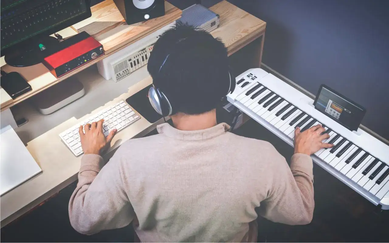 Mann mit Kopfh&ouml;rern spielt auf einem wei&szlig;en Keyboard und schreibt eigene Songs. Am Schreibtisch sind Computer, Lautsprecher und ein rotes Audio-Interface.