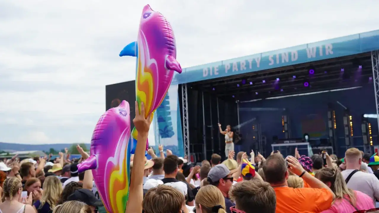 Bunte Delfin-Lufttiere fliegen über einer jubelnden Menge bei einem Festival. Die Bühne im Hintergrund zeigt die Aufschrift