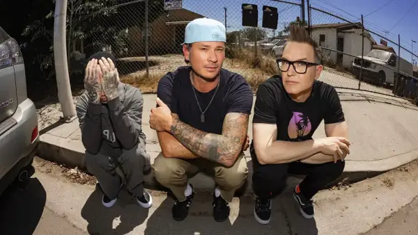 Die Mitglieder von blink-182 hocken auf dem Bürgersteig. Links Travis Barker, der sein Gesicht verdeckt. In der Mitte Mark Hoppus mit Cap und tätowierten Armen. Rechts Tom DeLonge mit Brille und Irokesenschnitt.