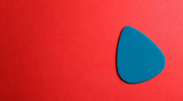 Ein blaues Plektrum liegt auf rotem Untergrund. Was ist ein Plektrum? Ein kleines Werkzeug zum Zupfen von Saiten.