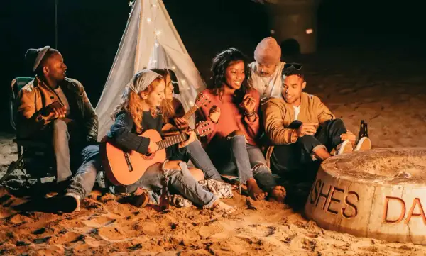 Freunde sitzen am Strand um ein Feuer, spielen Gitarre und singen Lagerfeuerlieder zum Mitsingen. Ein Zelt mit Lichterkette im Hintergrund.