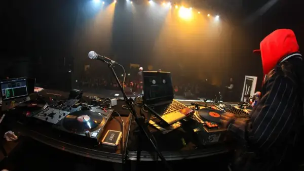DJ-Pult mit Plattenspielern, Laptop und Mikrofon. Im Hintergrund eine Bühne mit Scheinwerfern und Publikum. Die DJ Namen sind hier nicht sichtbar.