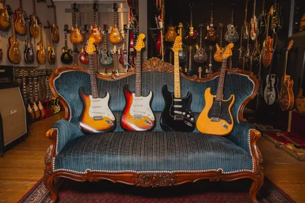 Vier E-Gitarren in verschiedenen gitarren formen und Farben ruhen auf einer antiken Couch, umgeben von einer Wand voller weiterer Gitarren.